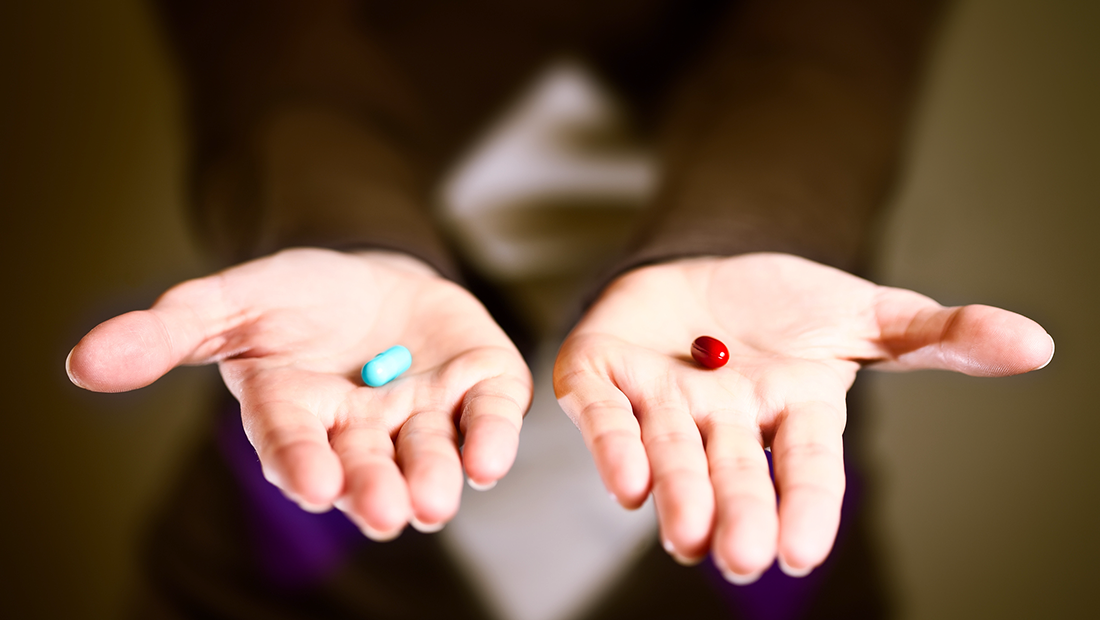 A blue medicine capsule in one palm and a red medicine capsule in the other.