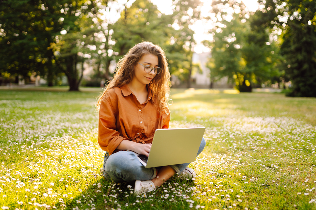 Naisoletettu istuu kesäisellä kukkaniityllä kannettava tietokone sylissään.