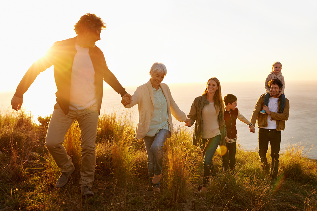 Joukko eri-ikäisiä nauravia ihmisiä kulkevat käsikädessä vuoren rinnettä ilta-auringossa.